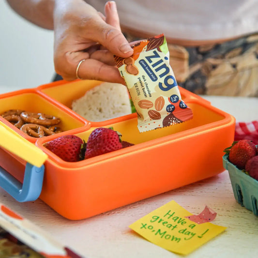 placing a zing bar mini into a lunch box with a sandwich, pretzels, and strawberries 